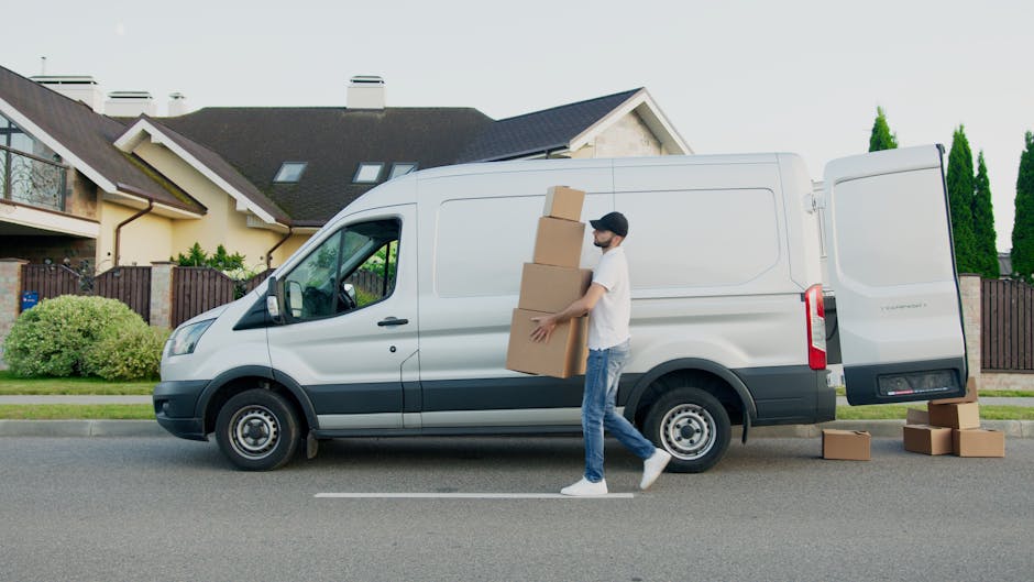 A man wearing a white polo shirt and a black cap is standing indoors next to a stack of uniformly sized cardboard boxes, which are arranged in a neat pile against a wall. He is holding a clipboard and