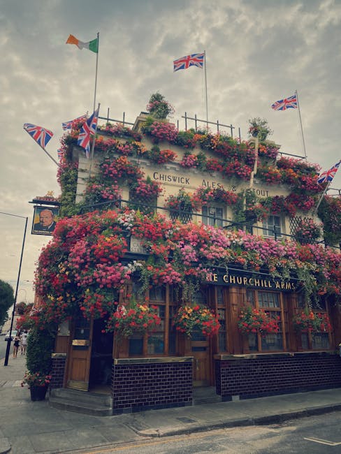The exterior of a traditional pub building in Chiswick, decorated with abundant hanging flower baskets and climbing plants covering the facade, including pink and red flowers. The pub features wooden window frames and a sign reading 'The Churchill Arms.' Multiple Union Jack flags and an Irish flag are mounted on flagpoles atop the building, fluttering in the breeze under a partly cloudy sky. The street view shows a pavement and a small black sign with a portrait and text, alongside a black bollard. The image evokes a lively atmosphere typical of a popular local pub, suitable for house removals or relocation services that include moving or packing-related activities in a historic or residential area like Chiswick.