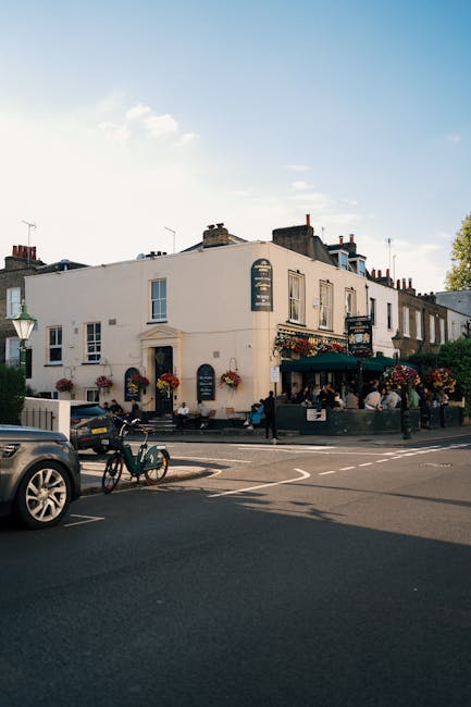 A two-storey white building situated on a street corner, hosting a pub or restaurant with outdoor seating. The building features large windows, decorative flower boxes beneath some of them, and a dark green awning above the entrance. Several hanging flower baskets adorn the facade, adding colour to the exterior. In front of the building, on the pavement, there are a few pedestrians, and a group appears to be gathered near the entrance or outdoor seating area. Parked along the road are a black car and a bicycle secured to a bike rack. The scene is illuminated by daylight with a partly cloudy sky overhead, and the surroundings include other buildings with chimneys in the background. This image captures a typical street scene in Chiswick, with potential relevance to house removals or moving services provided by Man and a Van Chiswick, showcasing an active urban environment and urban moving logistics.