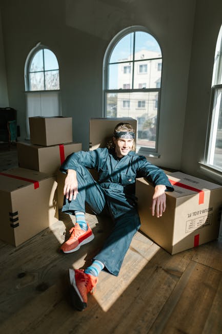 A young man in casual blue clothing and bright orange sneakers is sitting on the wooden floor inside a room with large arched windows that let in natural daylight. He is surrounded by several cardboard boxes, some sealed with red and black packing tape, indicating packing for a home relocation. The boxes are stacked and arranged around him, with some resting against the windowsill and others on the floor. The room has a neutral coloured wall, and the background reveals a view of neighboring buildings through the windows. The scene captures the loading and packing process involved in moving house, with the presence of furniture and packaging materials suggesting a professional moving operation. As part of [COMPANY_NAME]'s removals services, this image depicts the typical steps of furniture transport and packing during a same-day move in the W4 area, reflecting efficient and careful handling of belongings for a house relocation.
